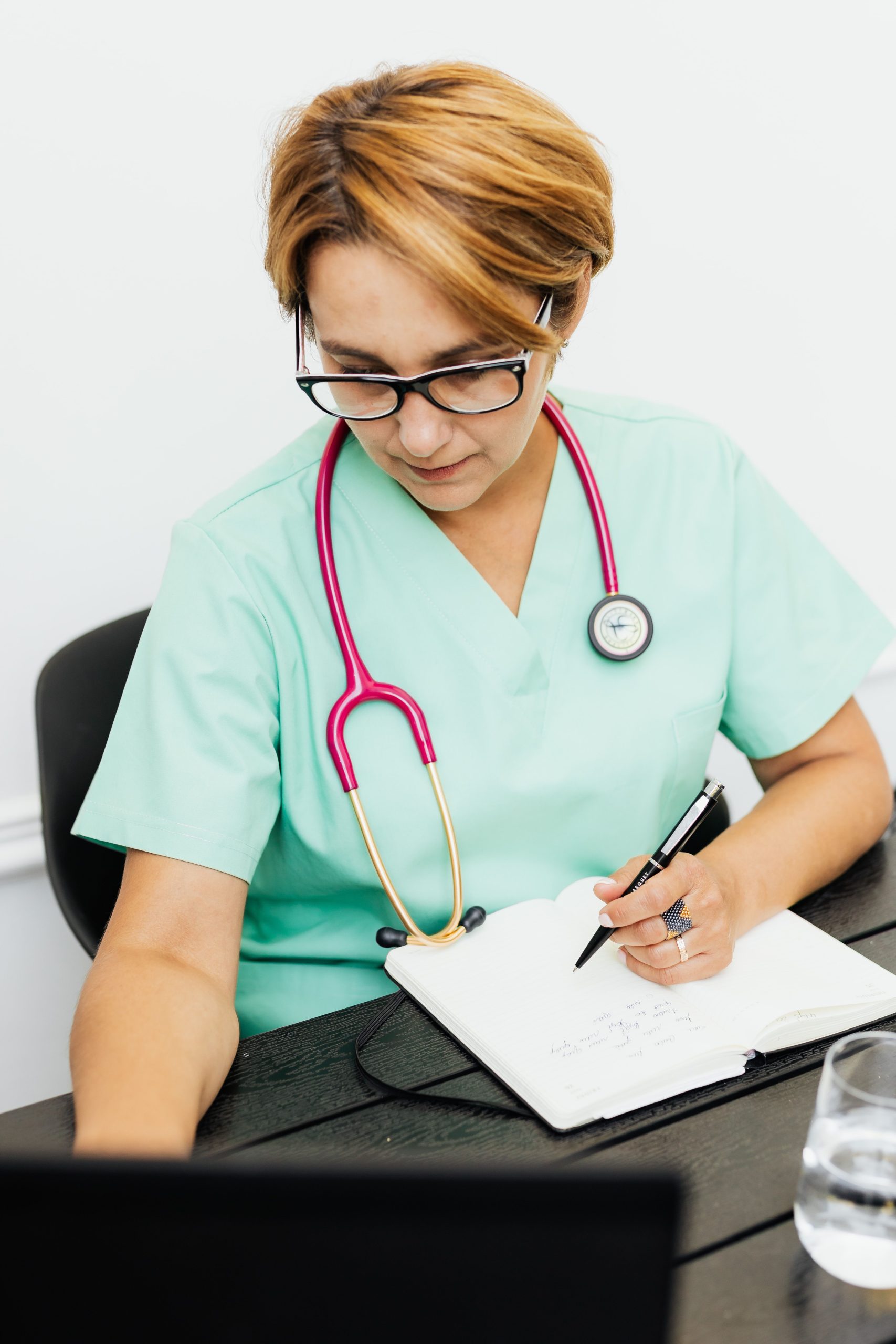 A female healthcare professional writing in a notebook with a stethoscope in a medical setting.